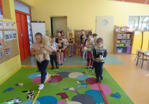 Dzieci ustawione w dwóch rzędach, trzymając misie pluszowe w ręku przechodzi po ułożonych wzdłuż szarfach.
